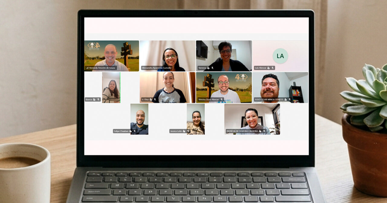 Notebook sobre uma mesa exibe uma videoconferência com diversos participantes em uma reunião on-line. Ao lado do computador, há uma xícara e um vaso com planta, compondo o ambiente de trabalho.