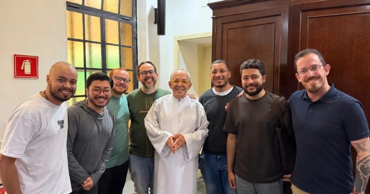 Participantes do Grupo de Acompanhamento Vocacional Inaciano (GAVI) posam para foto durante encontro no Centro MAGIS Anchietanum, em São Paulo (SP). O grupo reúne oito homens sorridentes, com um religioso de túnica branca ao centro, em ambiente interno.