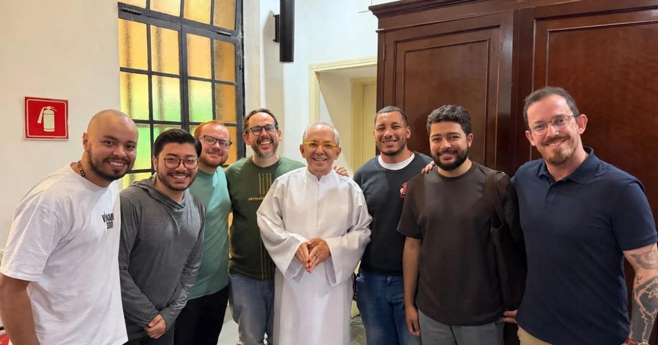 Participantes do Grupo de Acompanhamento Vocacional Inaciano (GAVI) posam para foto durante encontro no Centro MAGIS Anchietanum, em São Paulo (SP). O grupo reúne oito homens sorridentes, com um religioso de túnica branca ao centro, em ambiente interno.